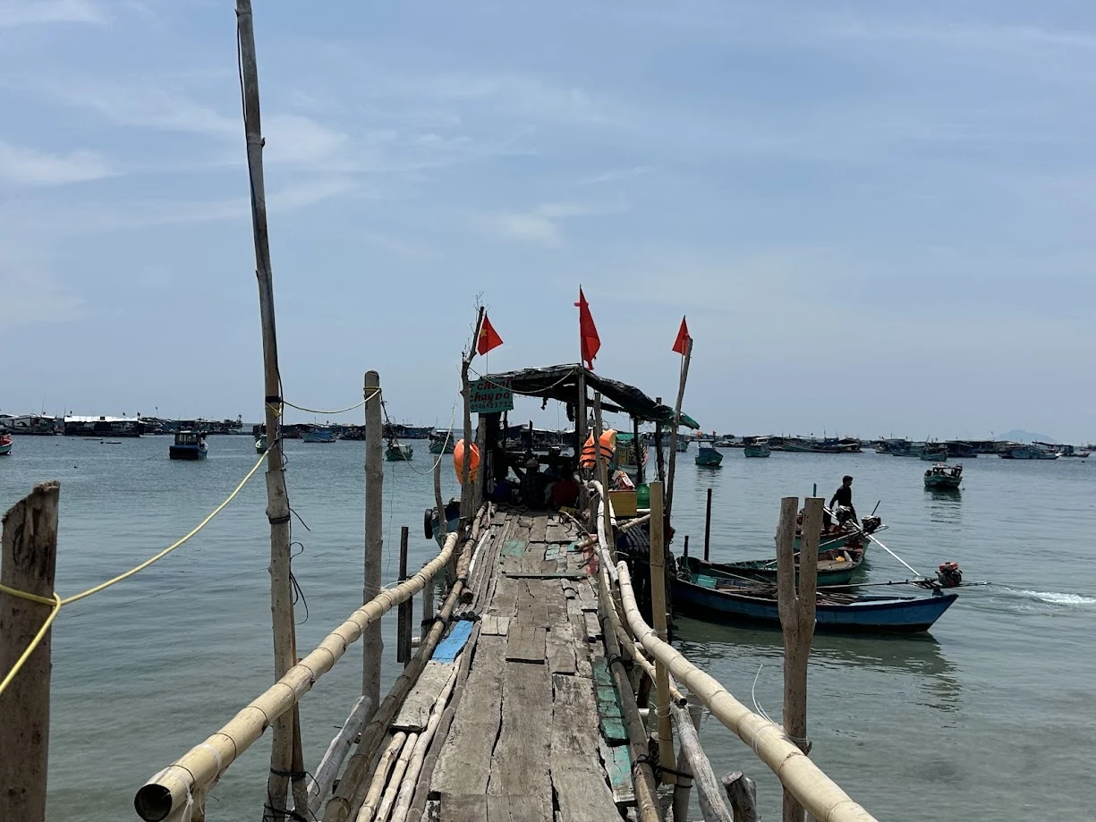 Cầu gỗ dẫn ra ghe tại làng bè Hòn Sơn, hai bên là tay vịn tre, phía xa là cụm bè neo trên biển