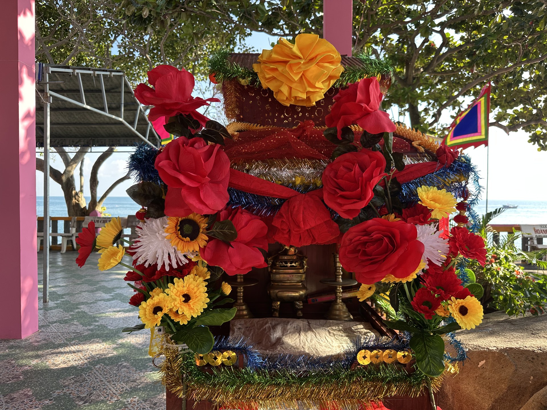 Một bàn thờ kết đầy hoa vải đỏ và vàng (hồng, cúc, hướng dương giả) cùng dây kim tuyến, đặt cạnh dãy mái hiên sơn hồng, dưới tán cây, nhìn ra biển