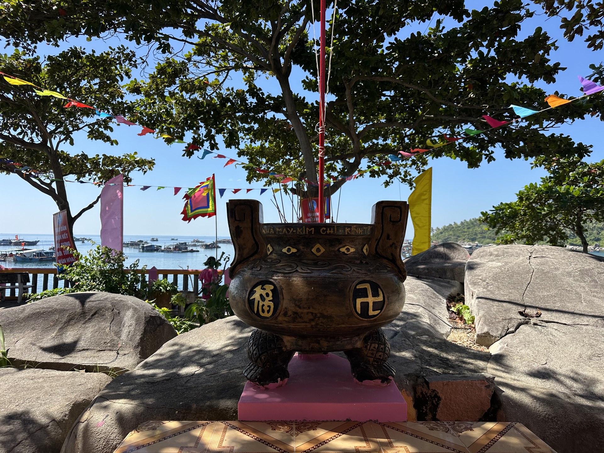 Lư hương đồng đặt trên tảng đá dưới tán cây trong khuôn viên lăng ở Hòn Sơn, dây cờ đuôi nheo nhiều màu giăng ngang, phía xa là ghe và bè neo trên mặt biển