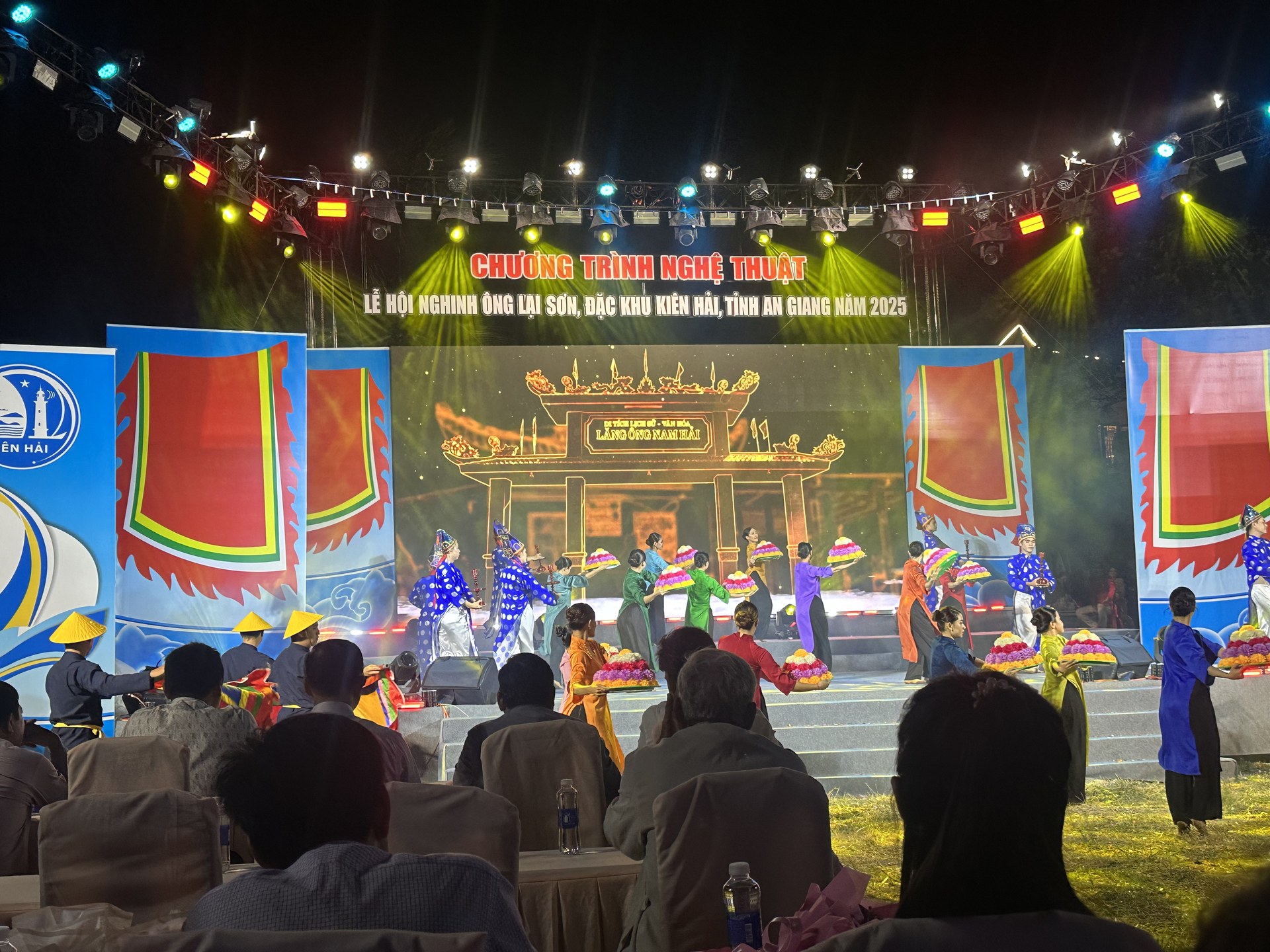 Sân khấu ngoài trời buổi tối ở Hòn Sơn, băng chữ ghi chương trình nghệ thuật lễ hội Nghinh Ông Lại Sơn năm 2025, diễn viên mặc áo dài nhiều màu múa dâng mâm hoa, khán giả ngồi xem phía dưới
