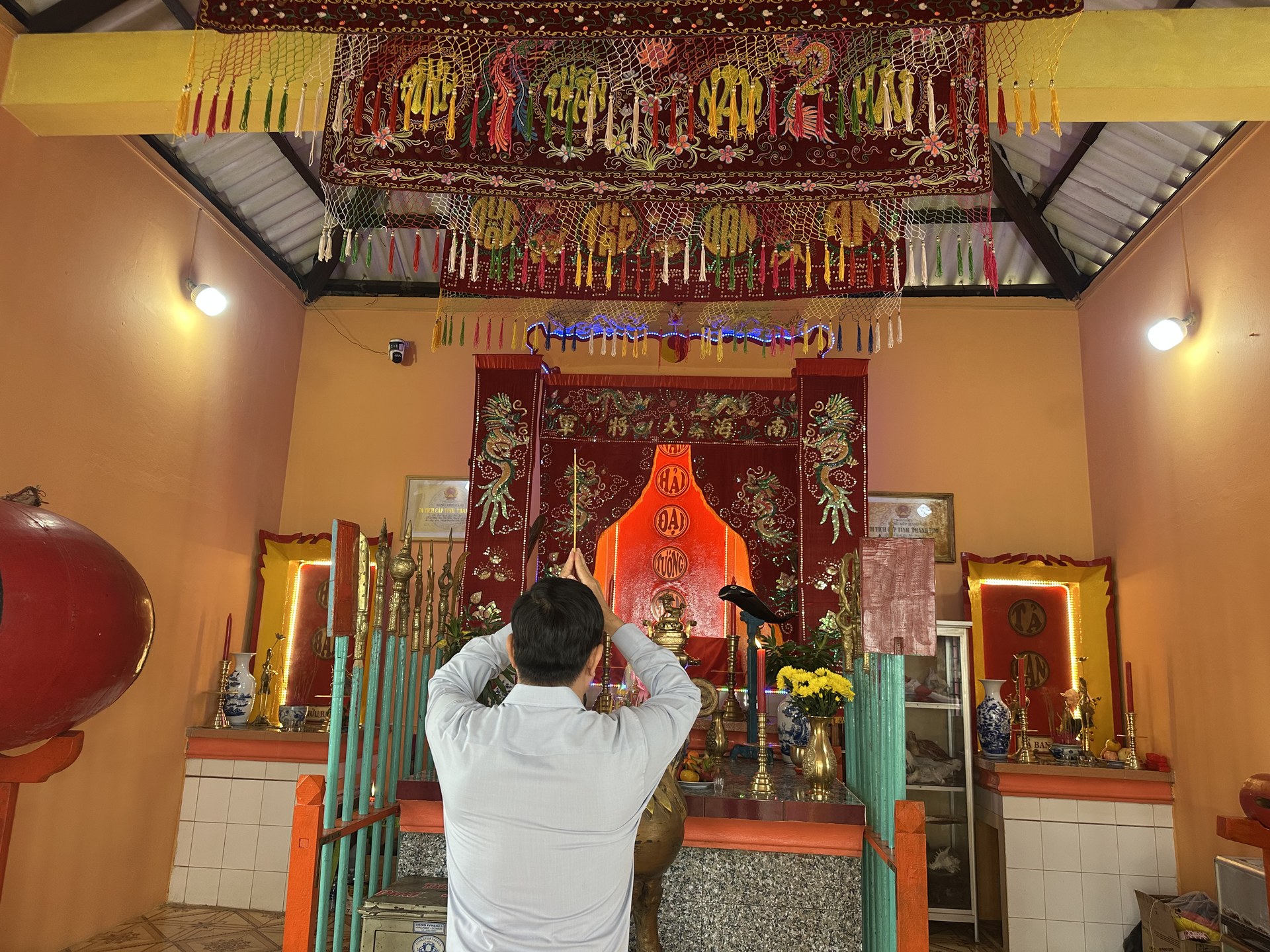 Một người quay lưng lại ống kính, hai tay nâng nhang trước bàn thờ chính thờ Nam Hải Đại Tướng Quân trong lăng ở Hòn Sơn, trần nhà rủ màn thờ thêu đỏ có tua